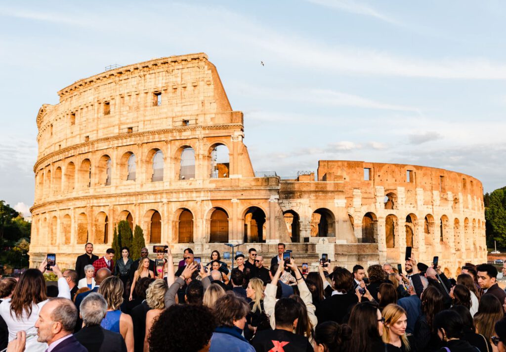 The cast of Fast X pose for a photo on a red carpet in front of the Colosseum in Rome, with others taking pictures and the sky clear in the background.