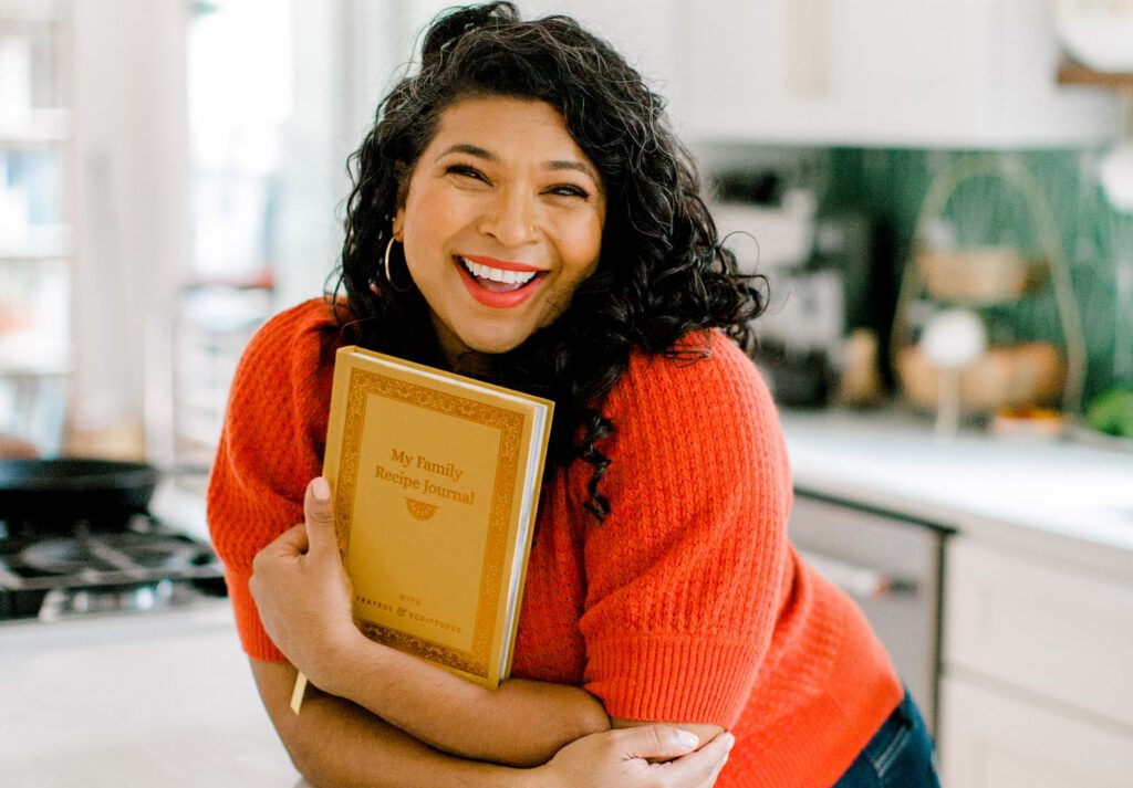 A smiling woman with curly hair, wearing a red sweater, stands in a bright kitchen while holding a yellow cookbook titled My Family Recipe Journal.