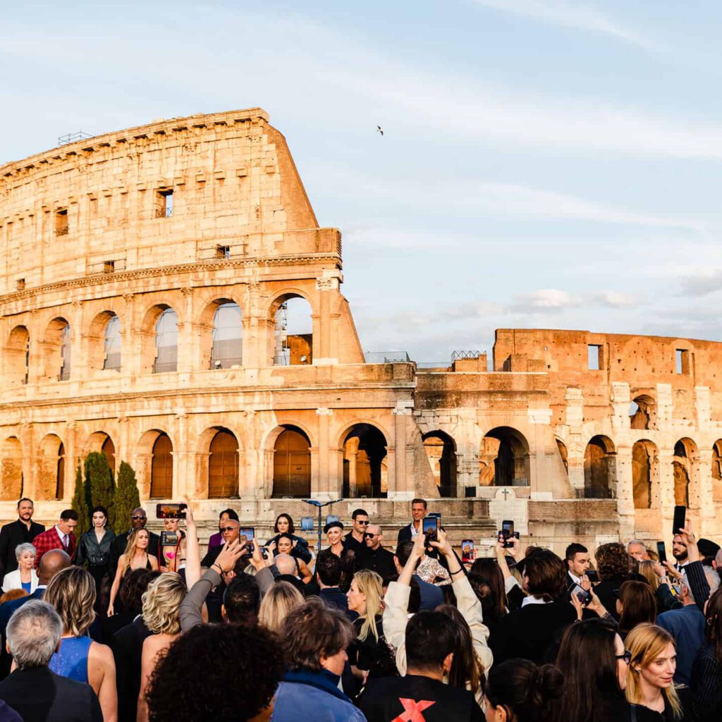 The cast of Fast X pose for a photo on a red carpet in front of the Colosseum in Rome, with others taking pictures and the sky clear in the background.