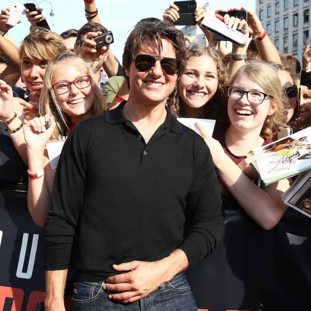 Tom Cruise wearing sunglasses and a black shirt smiles and poses with several excited fans holding photos and pens at an outdoor event. The crowd is gathered behind a barrier, and everyone looks happy.