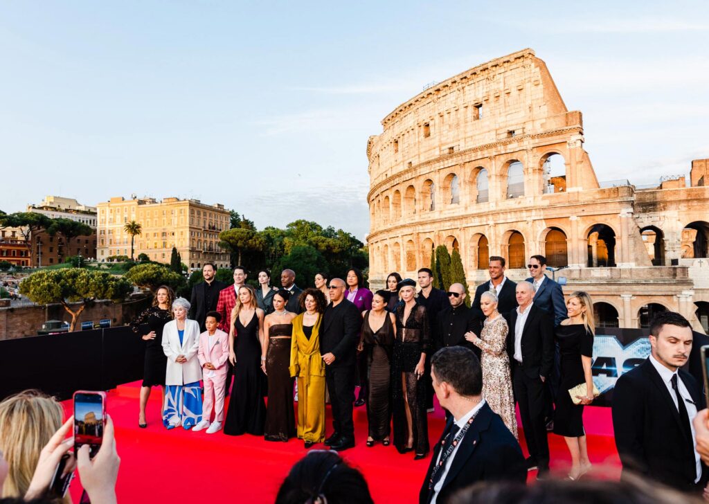 The cast of Fast X pose for a photo on a red carpet in front of the Colosseum in Rome, with others taking pictures and the sky clear in the background.