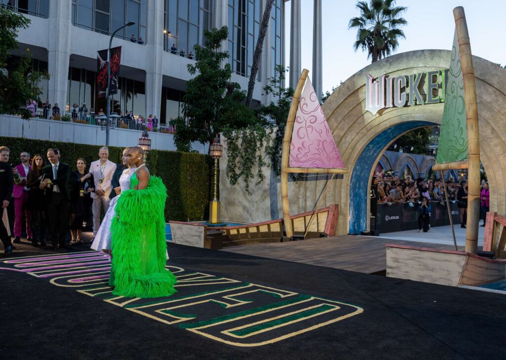 Ariana Grande and Cynthia Erivo pose on the wicked red carpet; one wears a shiny pale dress with gloves, the other wears a bright green dress with a dramatic fringe cape. People stand in the background near trees and a building.