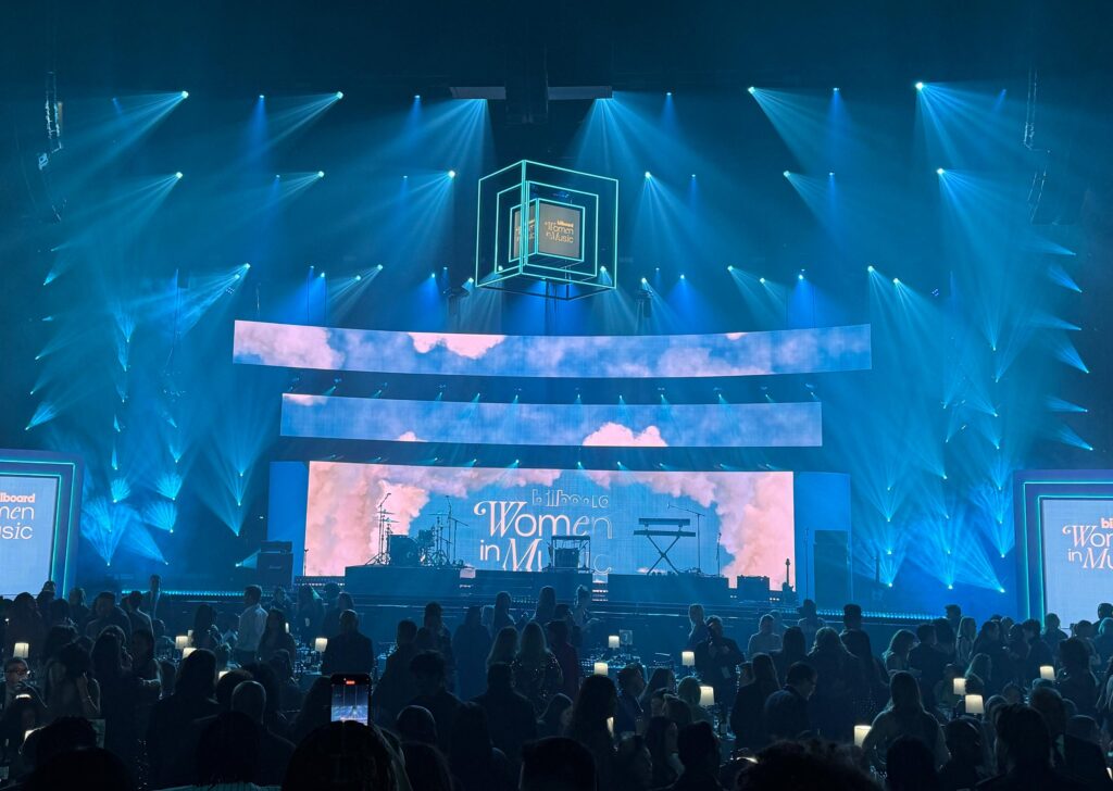 A large stage at the Billboard Women in Music event is lit with blue lights and digital screens, with musical instruments set up and an audience seated at tables in the foreground.