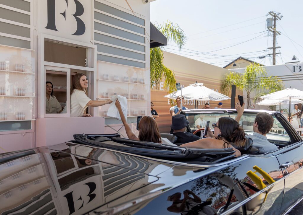 A woman hands a bag to people in a convertible at a Fenty Beauty drive-thru window, while others wait in cars behind them. The scene is sunny, with umbrellas, palm trees, and a striped building in the background.