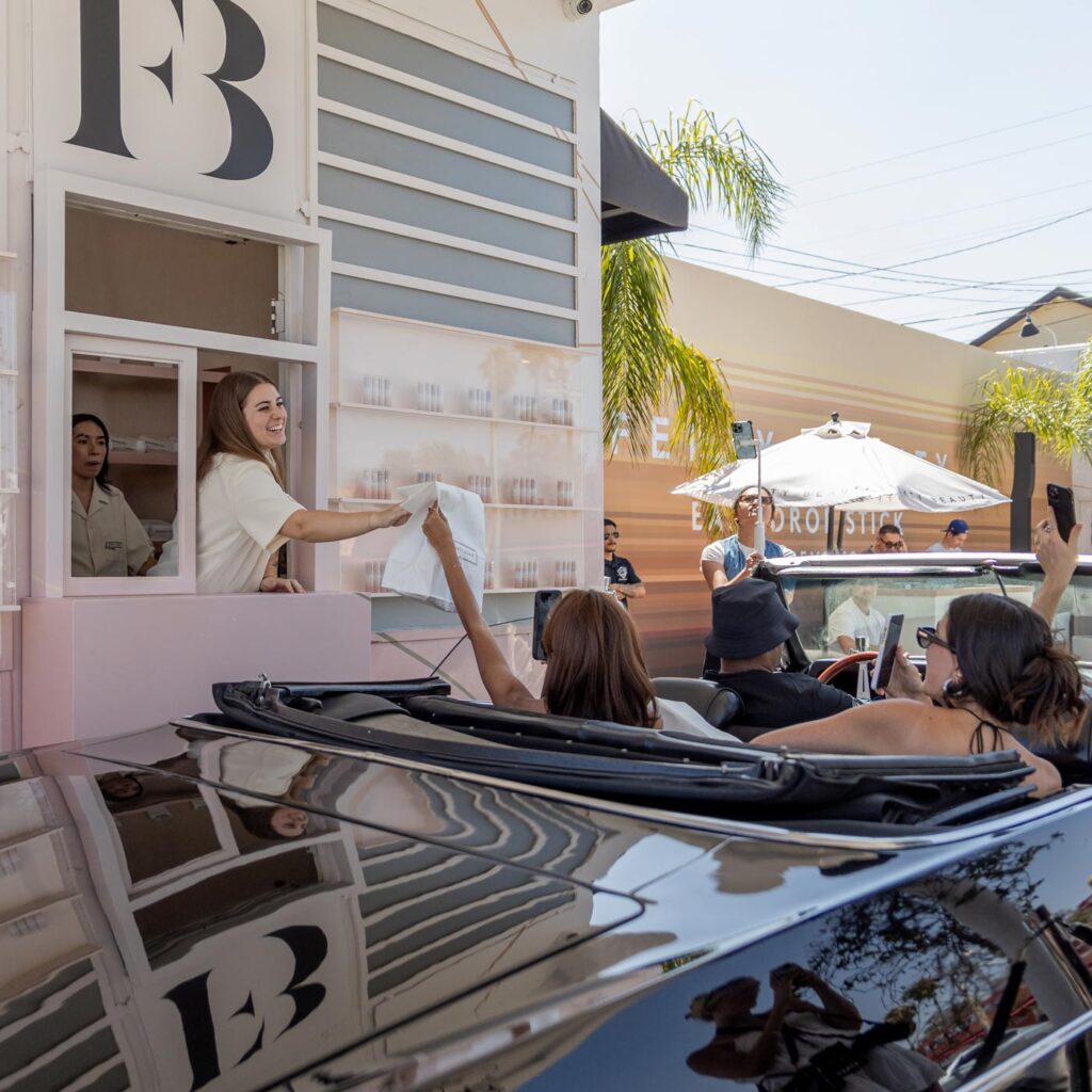 A woman hands a bag to people in a convertible at a Fenty Beauty drive-thru window, while others wait in cars behind them. The scene is sunny, with umbrellas, palm trees, and a striped building in the background.