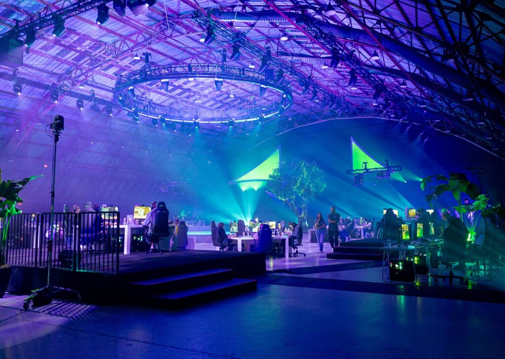 A large indoor event space with purple and blue lighting, filled with people seated at tables and computer stations, with a stage, plants, and modern decor under a high arched ceiling.