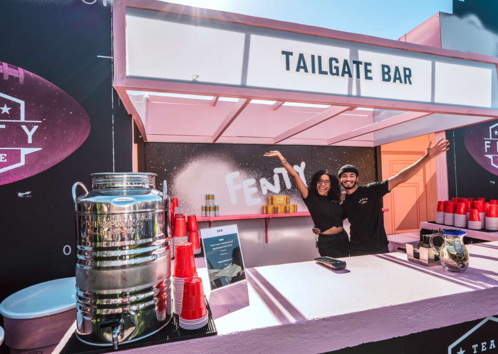 Two smiling people behind the counter of a Fenty Beauty Event Tailgate Bar stand with their arms raised. The counter displays red cups, a drink dispenser, and a menu. The background features a Fenty sign and vibrant decor.