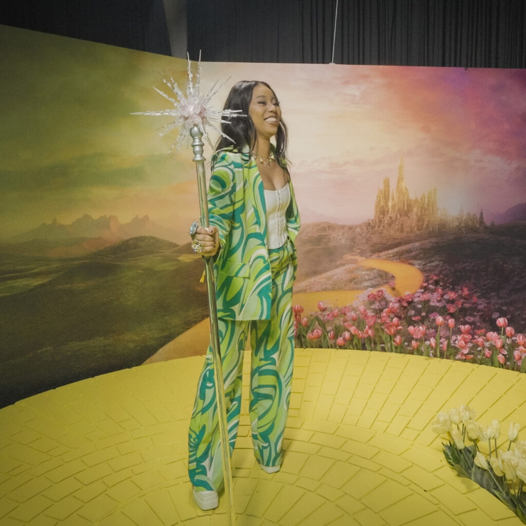 Girl in green pantsuit holding Glinda's wand and smiling big while standing on the yellow brick road