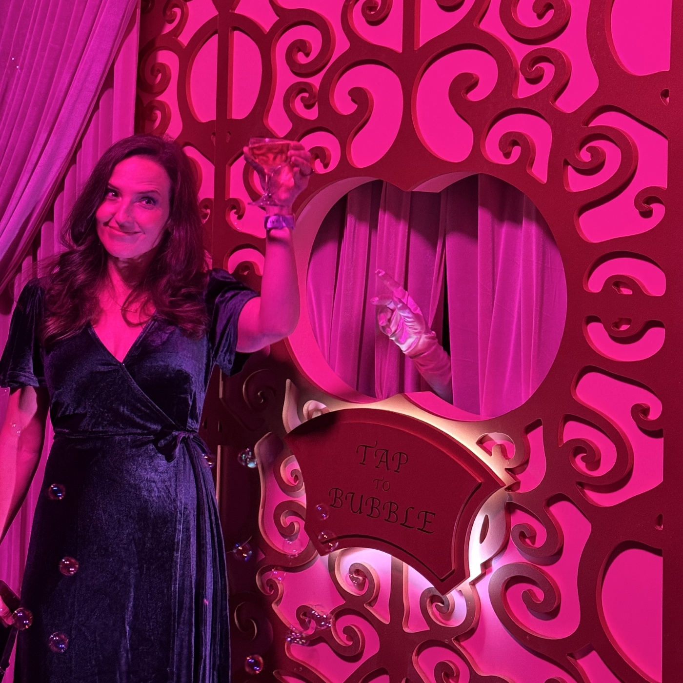 Girl standing in front of a decorated pink wall, holding a glass of champaign in her hand. There is a sign that reads "tap for bubbles" just below a whole in the wall from which a hand with a pink silk glove reaches out.