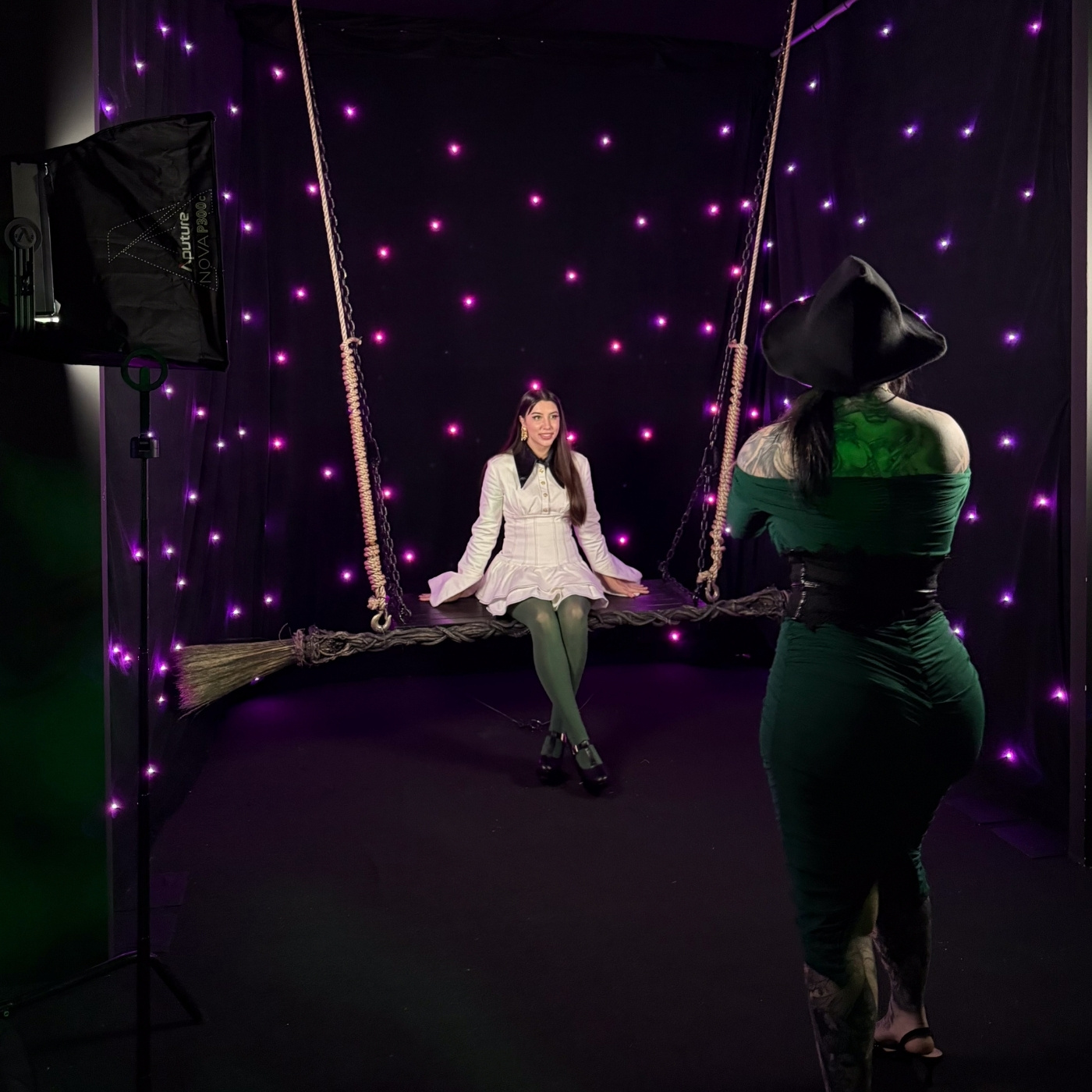 Girl dressed in white dress sits on a hanging broom smiling to the camera. Another girls dressed in an Elphaba outfit takes a photo of the girl on the broom swing. The background is black with twinkling purple neon lights.