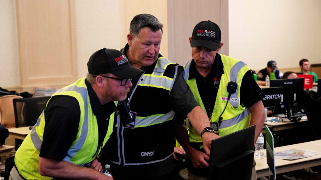 Three men dressed in safety gear and wearing Redrock Safety-branded hats are looking at a computer screen inside a command center.